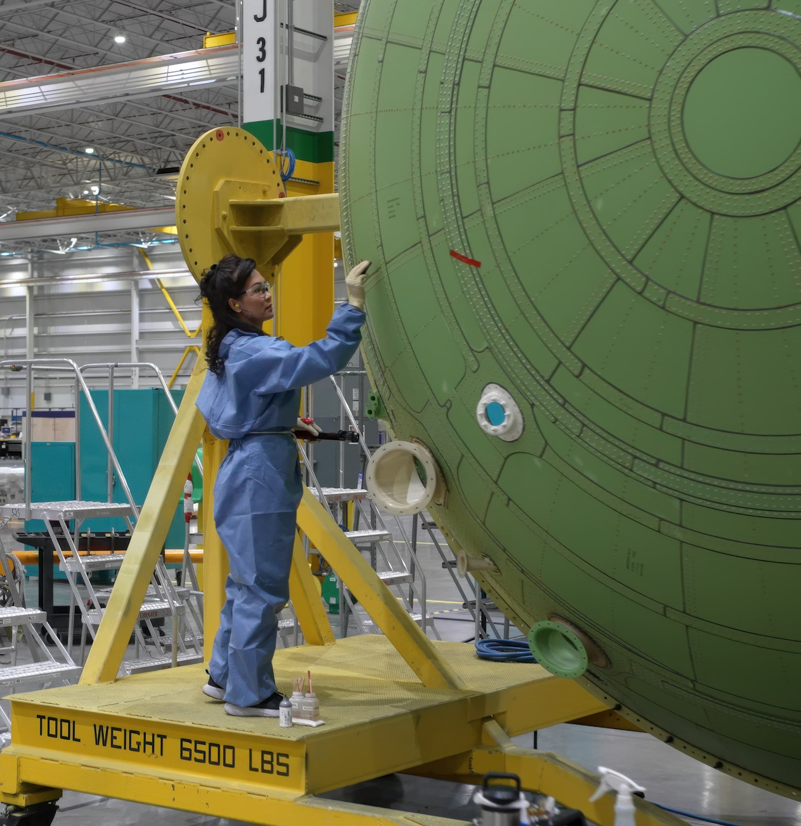 A technician in blue coveralls working on a green-primed fuselage barrel from a raised platform